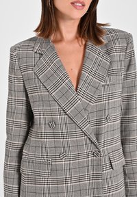 Blazer a quadri grigio con design doppiopetto, revers affilati, spalle strutturate e due tasche frontali con dettagli a bottone.