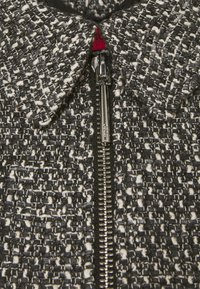 Chaqueta de tweed en blanco y negro con cuello, que presenta una cremallera plateada y una textura tejida intrincada. Los detalles incluyen una etiqueta roja.