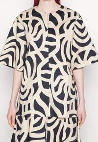 Top beige con motivi astratti neri, maniche corte e uno spacco frontale. Realizzato in tessuto liscio con un taglio ampio e confortevole.