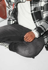 Czarna i biała koszula w kratę flanelową na białym swetrze, połączona z ciemnymi dżinsami i czarno-białymi sneakerami. Widać pozycję siedzącą.