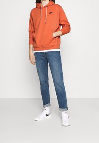 Orange Nike-hoodie med framficka, justerbar dragsko och svart logotyp. I kombination med uppvikta blå jeans och vita sneakers.