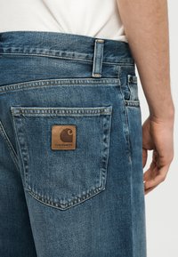 Κοντινό πλάνο ενός ατόμου που φοράει μπλε τζιν τζιν με ορατό το λογότυπο Carhartt στο πίσω τσεπάκι και μια λευκή πουκάμισα που έχει μπει μέσα.