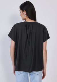 Sort kortærmet bluse lavet af blødt stof, med en rund halsudskæring og en plissédetalje på bagsiden. Sammen med lyseblå jeans.