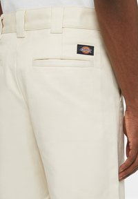 Pantaloni in twill di cotone beige con una tasca posteriore, cuciture rinforzate e un'etichetta nera e rossa con il logo Dickies sul lato destro.