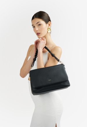 Donna con un vestito bianco che tiene una borsa nera in pelle con manici intrecciati e grandi orecchini a forma di fiore dorati su uno sfondo semplice.