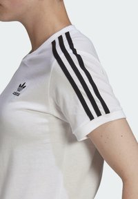 Balta medvilnės marškinėliai su juoda trijų juostų dizainu ant pečių, raštuota apykaklė ir mažas juodas logotipas viršutinėje krūtinės dalyje. Trumpi rankovės.