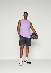 Lila ärmlös tröja med broderad text, svarta nätshorts och svarta sneakers med vita detaljer; håller i en basketboll.