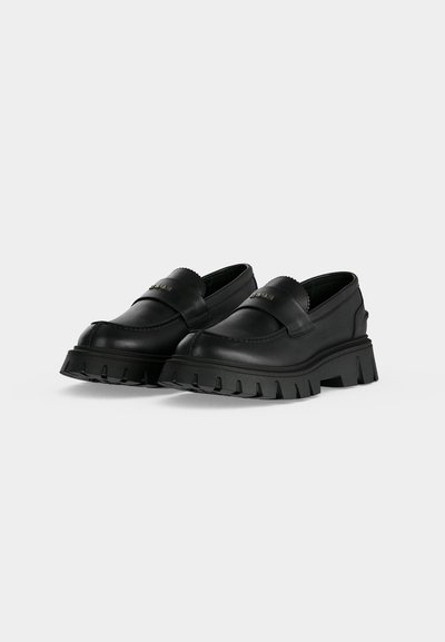 Mocassins noirs à semelle épaisse, dessus en cuir lisse et petit détail de logo métallique sur la bride, présentés sur un fond clair.