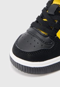 Sneaker nera con punta in pelle, pannelli in rete, accenti gialli, lacci rotondi neri e suola in gomma testurizzata con scanalature.