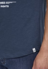 T-maica v mornarsko modri barvi iz mehkega blaga, z belim grafičnim besedilom in majhno etiketko na stranskem robu. Ujemajoče temne kavbojke.