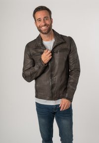Giacca di pelle marrone con chiusura a zip, caratterizzata da un colletto e una finitura testurizzata. Indossata sopra una maglietta grigia e abbinata a jeans blu.