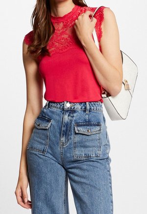 Vrouw draagt een mouwloze rode kanten top, hooggetailleerde blauwe spijkerbroek met klepzakken, draagt een witte handtas op haar schouder.