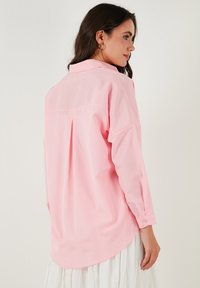 Light pink, long-sleeve shirt with a collared neckline, back pleat detail, and rounded hem. Fabric appears smooth and breathable.