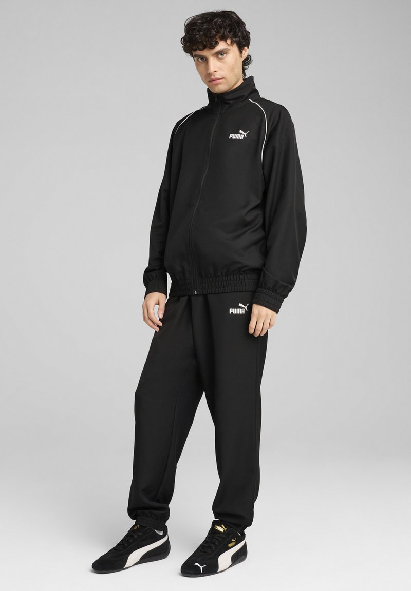 Black Puma tracksuit featuring a zip-up jacket with a high collar and elastic cuffs, paired with matching pants and black sneakers with white accents.