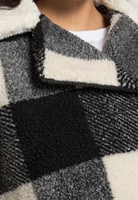 Eine Person trägt ein weißes Hemd unter einer Reißverschluss-Fleecejacke mit großen, schwarz-weißen und grauen Farbblockabschnitten sowie einem strukturierten Stoff.