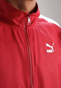 Chaqueta roja con cremallera hecha de material suave y ligero. Presenta un cuello alto, acentos blancos en las mangas y un logo blanco de Puma en el pecho.