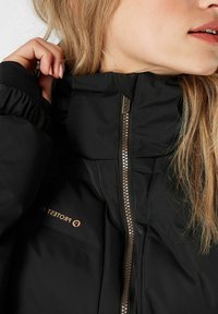 Giacca a vento nera con colletto alto, chiusura con zip dorata e polsini elastici. Il materiale ha una texture liscia e lucida con il marchio visibile.