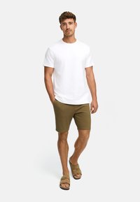 Homme marchant les mains dans les poches portant un t-shirt blanc uni, un short vert olive et des sandales beiges sur un fond blanc.