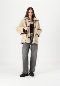 Beige fleece jack met bruine accenten, grote kraag en knoopsluiting, gecombineerd met grijs gestreepte wijde broek en zwarte schoenen.