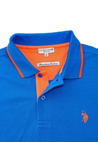 Modra polo majica z oranžnim ovratnikom in obrobo, dvema gumboma, in vezenem logotipom. Narejena iz vrhunskega bombaža, z teksturirano tkanino.