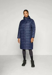 Mörkblå pufferjacka med huva, knälång, med horisontell sömnad, dragkedja fram och sidofickor. Bärs över svarta leggings och stövlar.