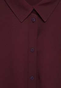 Gros plan d’un col de chemise bordeaux foncé avec une patte de boutonnage présentant trois boutons ronds visibles.