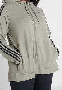 Hoodie zippé vert clair avec capuche à cordon, bandes latérales noires, et poches avant, fabriqué dans un tissu doux et lisse.