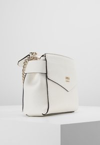 Borsa a tracolla in pelle bianca testurizzata con chiusura a patta, bordi neri e tracolla in metallo dorato, posata su un pedestal bianco contro uno sfondo grigio.