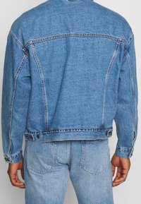 Blue denim jacket with a relaxed fit, featuring a classic back yoke, visible stitching, buttoned cuffs, and a slightly distressed texture.
