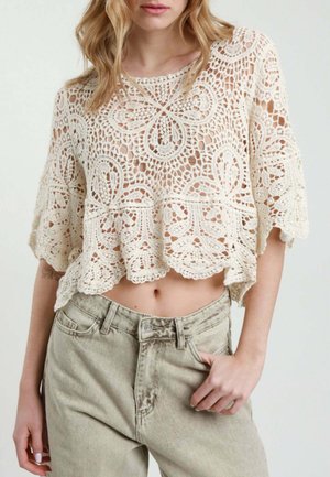 Donna che indossa un top corto in pizzo crochet crema con maniche corte e jeans beige chiaro a vita alta, mano poggiata nella tasca.