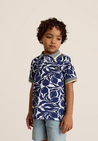 Polo shirt in marineblauw en wit met een bladerenpatroon, gestructureerde stof, en een grijze kraag met gele accenten, gecombineerd met lichtblauwe shorts.