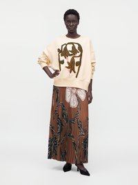 Femme portant un sweat crème avec broderie florale marron et une jupe plissée fleurie marron avec des talons noirs.