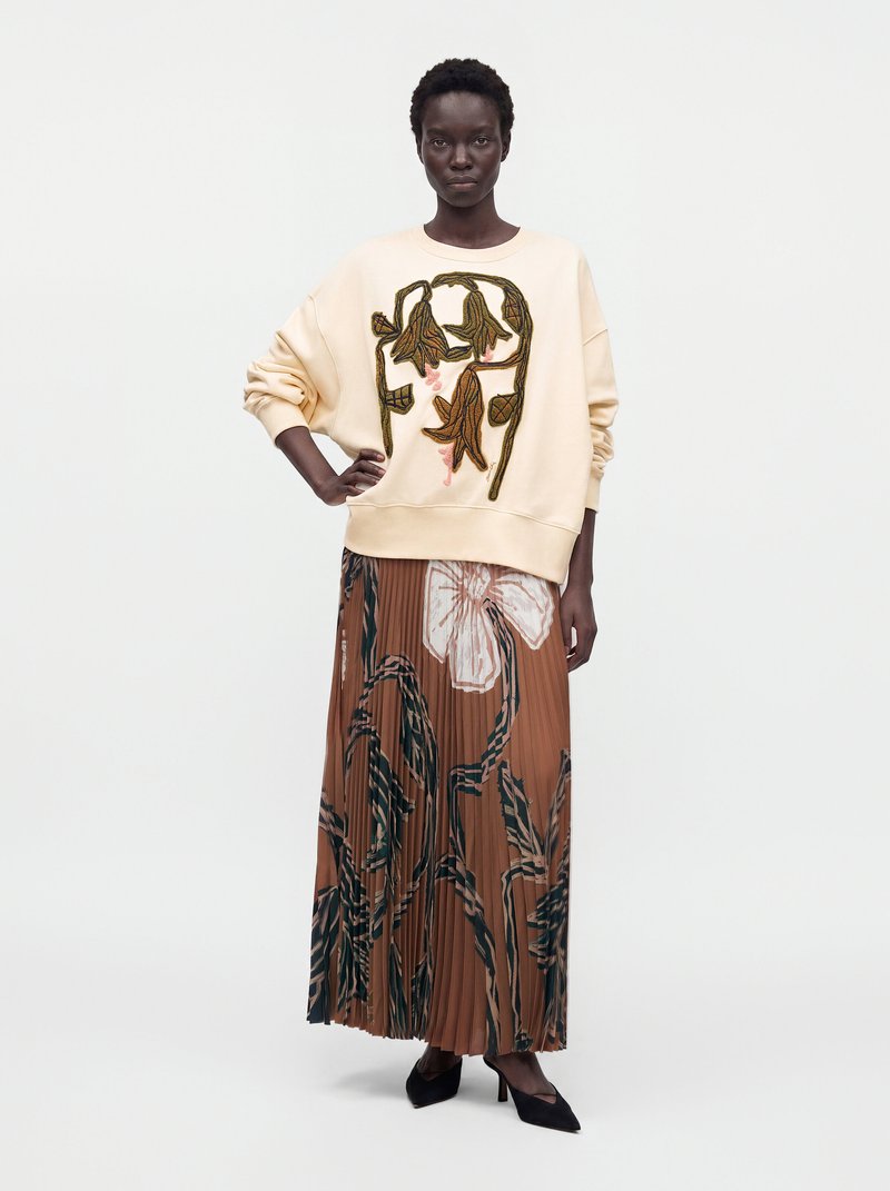 Femme portant un sweat crème avec broderie florale marron et une jupe plissée fleurie marron avec des talons noirs.