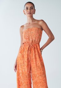 Oranje strapless jumpsuit met bloemmotief, geplooide buste, elastische taille en trekkoord. Loszittende, luchtige stof, twee zijzakken.
