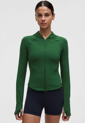 Vrouw draagt een nauwsluitende groene zip-up hoodie met duimgaten en zwarte sportshorts, staand tegen een effen lichte achtergrond.