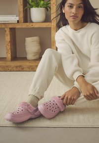 Roze klompen met een fuzzy roze voering, grote ventilatiegaten en een zachte textuur. Gedragen met lichtgekleurde joggers en sokken.