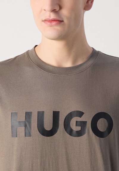 Сива памучна тениска с кръгло деколте и черно лого "HUGO", отпечатано на видно място отпред. Гладка текстура, минималистичен дизайн.