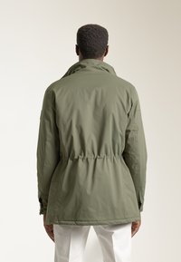 Chaqueta ligera de color verde olivo con cintura ajustable, cuello alto, y diseño minimalista. Textura suave y lisa, mangas largas.