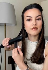 Ein schwarzer Haarglätter, der in einer Hand gehalten wird, glättet langes, glattes Haar. Der Benutzer trägt ein weißes, strukturiertes Oberteil und hat eine silberne Uhr am Handgelenk.