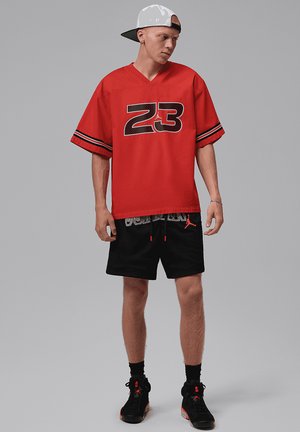 Ung mand iført en hvid kasket med kasketten bagud, rød sportstrøje med nummer 23, sorte basketballshorts og sorte høje sneakers mod en grå baggrund.