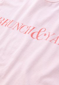 Svijetlo ružičasta pamučna majica s podebljanim crvenim tekstom "BRUNCH & YAY." Tkanina je meka i glatka, a kroj opušten.