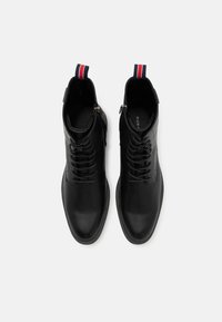 Botas de tobillo de cuero negro con una textura suave, punta redonda, cierre de cordones en la parte delantera y un tacón bajo, que presentan una lengüeta de tira de color en la parte trasera.