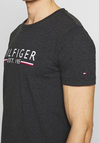 Camiseta de algodón gris con cuello redondo, que presenta el texto blanco "HILFIGER EST. 1985" y un pequeño logo rojo, blanco y azul en la manga.