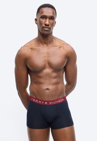 Μυώδης άνδρας που στέκεται με τα χέρια πίσω από την πλάτη, φορώντας μπλε σκούρα αντρικά εσώρουχα Tommy Hilfiger με κόκκινη μέση.