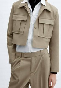 Veste beige cropped avec deux poches avant, assortie à une chemise blanche et un pantalon taille haute beige. Tissu lisse et design structuré.