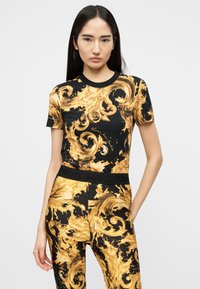 Γυναίκα που φοράει μαύρο και χρυσό τοπ με ριγωτό μπαρόκ σχέδιο και κοντά μανίκια, και παντελόνι με την ίδια εκτύπωση, με μέση "Versace Jeans Couture", στέκεται μπροστά σε μονόχρωμο φόντο.