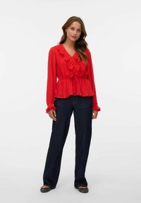 Blusa rossa a maniche lunghe con volant e vita legata, abbinata a pantaloni larghi in denim scuro e scarpe piatte nere e a fantasia.