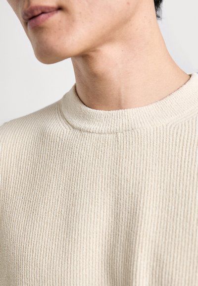 Pull beige côtelé avec un col montant et une surface texturée, mettant en avant un gros plan des détails du tissu et des coutures.