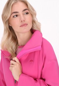 Rožinė vandeniui atspari striukė su aukštu apykakle, kuriame yra siuvinėjimas su skėčiu ir užtrauktukas. Po ja iš dalies matoma dryžuota medžiaga.