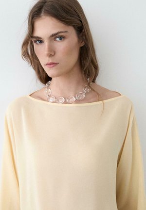 Femme aux cheveux châtain clair portant un haut jaune pâle épaules dénudées et un collier fait de grosses perles transparentes sur un fond clair uni.